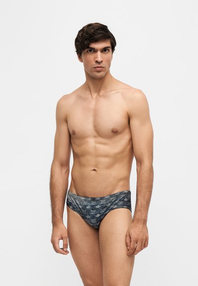 Jeune homme aux cheveux foncés se tient devant un fond uni, portant un slip de bain à motifs, regardant droit devant avec une expression neutre.