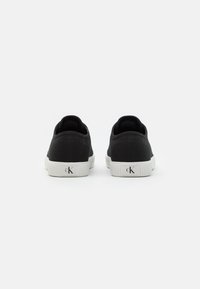 Ett par svarta låga sneakers med vita sulor, sedd från baksidan, med "cK"-logotypen på hälen mot en vit bakgrund.