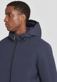 Giacca con cappuccio blu navy con chiusura a zip. Caratterizzata da un tessuto testurizzato, polsini elasticizzati e una toppa con logo sulla spalla sinistra.