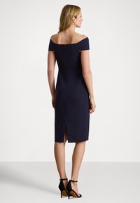 Μπλε σκούρο φόρεμα με ανοιχτούς ώμους, εφαρμοστή γραμμή, μήκος μέχρι το γόνατο και σκίσιμο στο πίσω μέρος. Διαθέτει κρυφό φερμουάρ και λείο ύφασμα.