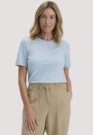 Frau mit schulterlangem blondem Haar, die ein hellblaues Kurzarmhemd und beige Kordelzughosen trägt, posiert mit einer Hand in der Tasche.