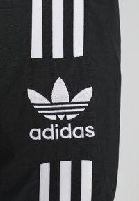Svart tyg med vit Adidas-logotyp och tre vertikala ränder i texturerat material. Logotypen har en bladdesign med horisontella linjer.