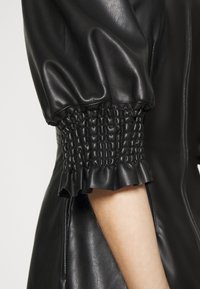 Manche de veste en similicuir noir avec poignet élastiqué et froncé et bordure à volants, exposant partiellement l'avant-bras sur un fond clair.