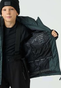 Zielona ocieplana kurtka z czarnym wnętrzem, z etykietą, siateczkową kieszenią i brandingiem SWEDEMOUNT. Noszona na czarnym prążkowanym swetrze.