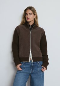 Veste bomber marron avec un corps en suédine et des manches en tricot côtelé, dotée d'une fermeture éclair à l'avant, de poches latérales et de textures contrastées. Portée avec un jean bleu.