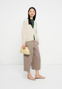 Cardigan bege claro sobre uma blusa verde escura, combinado com umas calças largas bege e sapatos baixos cor-de-rosa, segurando uma pequena mala amarela.