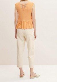 Woman wearing an orange sleeveless top with a ruffled hem and tie back, off-white cropped pants, and beige quilted slide sandals.