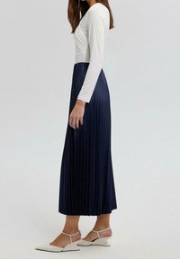Lunga gonna plissettata blu abbinata a un top fitted bianco a maniche lunghe. L'outfit include scarpe slingback bianche con un tacco angolare unico.