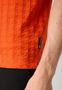 T-shirt laranja texturizado com um padrão geométrico, apresenta uma etiqueta lateral com o nome da marca. Exibido contra um fundo escuro.