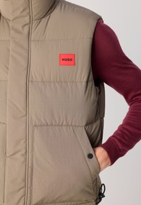 Taupe padded vest with a red HUGO logo patch worn over a burgundy long-sleeve sweater, hand in vest pocket, against a plain background.