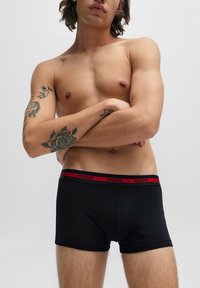 Schwarze Baumwoll-Boxershorts mit einem roten HUGO-Logo-Bund, die eine enganliegende Passform und eine glatte Textur bieten. Haut ist über dem Bund sichtbar.
