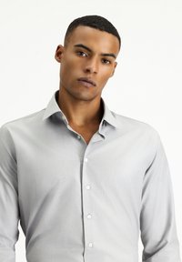 Lichtgrijze overhemden van een gladde stof, met een klassieke kraag, lange mouwen en een sluiting met knopen aan de voorkant met witte knopen.