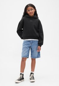 Meitene ar cirtainiem matiem, valkājot caurspīdīgas brilles, melnu kapuci ar GAP logo, gaišas džinsa šortus, baltas zeķes un melnas augstās teniskas.