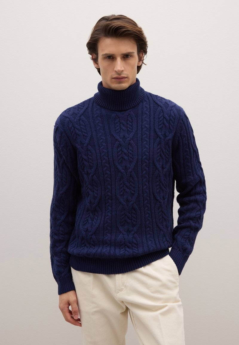 Navy blue cable-knit sweater with a high turtleneck and ribbed cuffs, featuring a textured pattern and a relaxed fit. Paired with light-colored pants.