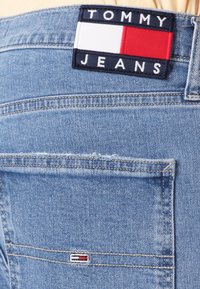Plave traperice s tkaninskom oznakom na kojoj piše "TOMMY JEANS" bijelim slovima na tamnoplavoj pozadini, uz crveno-bijeli pravokutni naglasak.