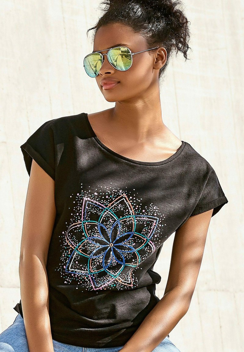 Camiseta negra de manga corta con mangas enrolladas, que presenta un diseño de mandala colorido con acentos en azul, rosa y blanco, sobre un fondo liso.