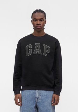 Jonge man met gevlochten haar, die een zwarte GAP-trui en blauwe spijkerbroek draagt, staand tegen een eenvoudige lichte achtergrond.