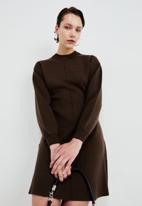Nainen, jolla on lyhyet tummat hiukset, korvissaan renkaat ja päällään tiukka pitkähihainen ruskea mekko, pitelee mustaa käsilaukkua valkoista taustaa vasten.