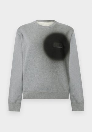 Bluză gri deschis cu gât rotund, mâneci lungi, manșete și tiv striate, și un patch circular negru pe partea stângă a pieptului cu o etichetă mică.