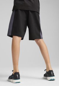 Pantaloni sportivi neri con pannelli laterali grigi, vita elastica, design fino al ginocchio, abbinati a sneakers nere con suola blu.