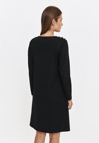 Czarna sukienka z długim rękawem o luźnym kroju, z złotymi guzikami na ramionach i miękkiego materiału, sięgająca do połowy uda.