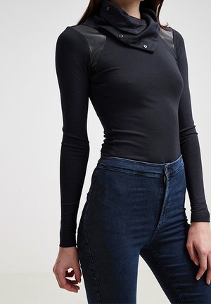 Nainen, joka pukeutunut mustaan, vartaloa myötäilevään pitkähihainen puseroon, jossa on korkea kaulus, ja tummansinisiin korkeavyötäröisiin farkkuihin, seisoo yksinkertaisen taustan edessä.