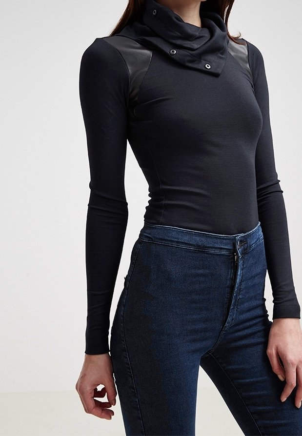 Woman wearing a fitted black long-sleeve top with a high collar and dark blue high-waisted jeans, standing against a plain background.