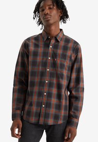 Camisa de cuadros en gris oscuro con rayas naranjas y azules. Diseño tipo botón, mangas largas y un bolsillo en el pecho con un pequeño logotipo.