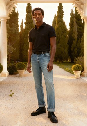 Jeune homme portant un polo noir, un jean bleu clair et des chaussures noires se tient sur un chemin de jardin ensoleillé avec de grands arbres et des colonnes derrière lui.