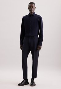 Camicia a maniche lunghe blu navy con chiusura a bottoni, abbinata a pantaloni sartoriali; entrambi realizzati in tessuto liscio. Scarpe nere stringate completano il look.