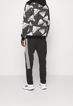 Personne portant un hoodie Adidas noir et blanc avec un motif de logo répétitif, un pantalon de survêtement Adidas noir avec des bandes blanches et des baskets Adidas blanches.