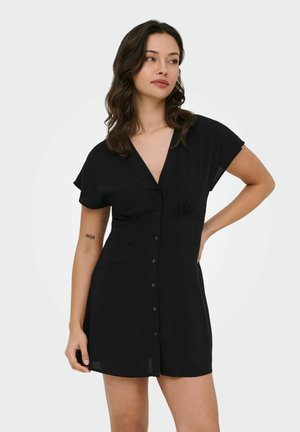 Kvinde med mørkt bølget hår iført en kort sort kjole med knapper, stående med den ene hånd på hoften og kiggede sidelæns.