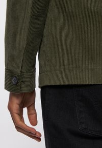 Kurtka z kordurowej tkaniny w kolorze oliwkowym, z poziomymi ribbkami, z mankietem na guziki i prostym dołem, w zestawie z czarnymi dżinsami.