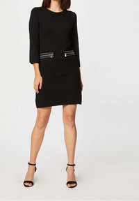 Robe noire à manches trois-quarts, avec deux poches zippées à l'avant et une coupe droite. Tissu lisse sans motif. Sandales noires à talons hauts.