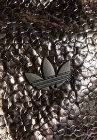 Metaliczna teksturowana powierzchnia w ciemnych odcieniach z wytłoczonym w szarości logiem trójlistnej koniczyny, z poziomymi liniami na środkowym listku.