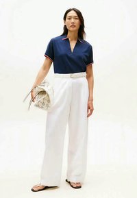 Navy polo shirt with red trim, white wide-leg trousers, white belt, and patterned handbag. Model wears black flip-flops.