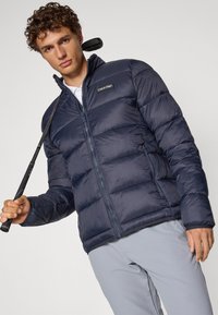 Marineblå puffjakke med glidelås foran og høy krage. Har et Calvin Klein-logo. Modellen holder en golfkjølle og har på seg lyse grå bukser.