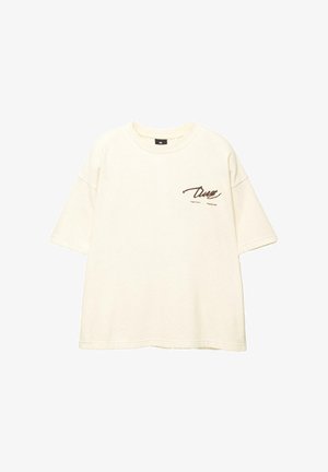 Valkoinen ylipitkä t-paita pehmeästä kankaasta, jossa on kalligrafinen logo vasemmassa rintataskussa ja lyhyet hihat.