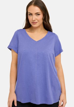 Vrouw met lang bruin haar die een lavendelkleurig T-shirt met korte mouwen en een V-hals draagt, terwijl ze voor een eenvoudige lichte achtergrond staat.