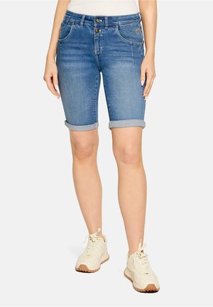 Vrouw draagt blauw denim shorts van middelmatige lengte met opgerolde pijpen en beige sneakers, staand tegen een effen achtergrond.