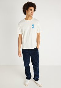 Tricou scurt, de culoare crem, din bumbac, cu un design grafic albastru pe piept, asortat cu pantaloni din corduroy bleumarin și adidași albi.