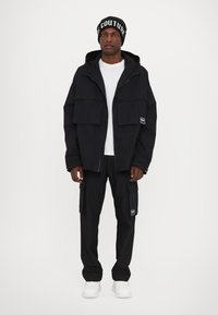 Black hooded jacket with two large chest pockets, paired with matching cargo pants and white sneakers. Features a beanie with text.