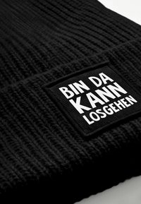 Black knitted beanie featuring a textured fabric and a rectangular patch with white bold text that reads "BIN DA KANN LOSGEHEN."