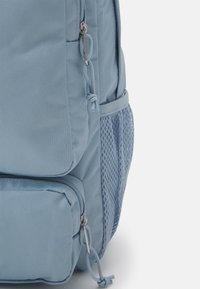 Mochila de color azul claro hecha de material texturizado, con un bolsillo grande, un bolsillo más pequeño con cremallera y un bolsillo lateral de malla.