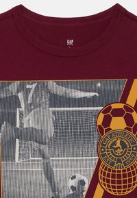 T-shirt pour enfant couleur bordeaux avec un graphisme d'un joueur de football frappant un ballon près du but et un emblème indiquant "GAP Athletic Est 1969" avec un ballon de football.