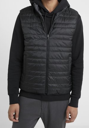 Mann trägt eine schwarze gesteppte ärmellose Weste über einem schwarzen Hoodie und grauen Hosen, steht vor einem einfarbigen weißen Hintergrund.