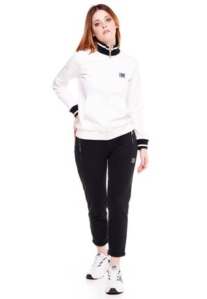 Une femme présente une veste blanche zippée avec des bordures noires, un pantalon noir et des baskets blanches sur un fond blanc uni.