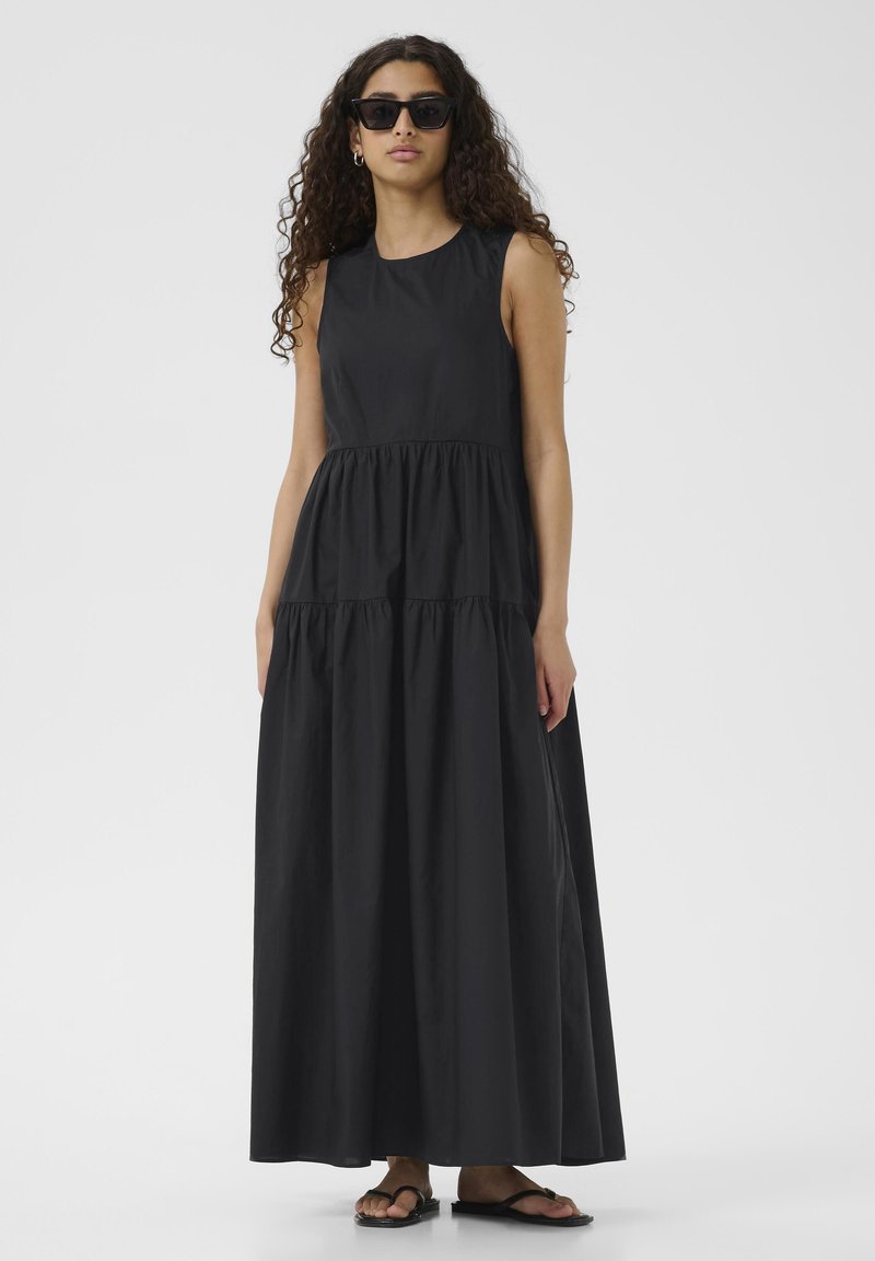 Schwarzes ärmelloses Maxikleid im gestuften Design mit glatter Textur. Das Modell trägt dazu schwarze Sonnenbrillen und Flip-Flops.