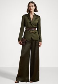 Olive green satin suit with a fitted blazer and wide-leg pants, accented by a brown belt with a chain detail. A small dark clutch is held.