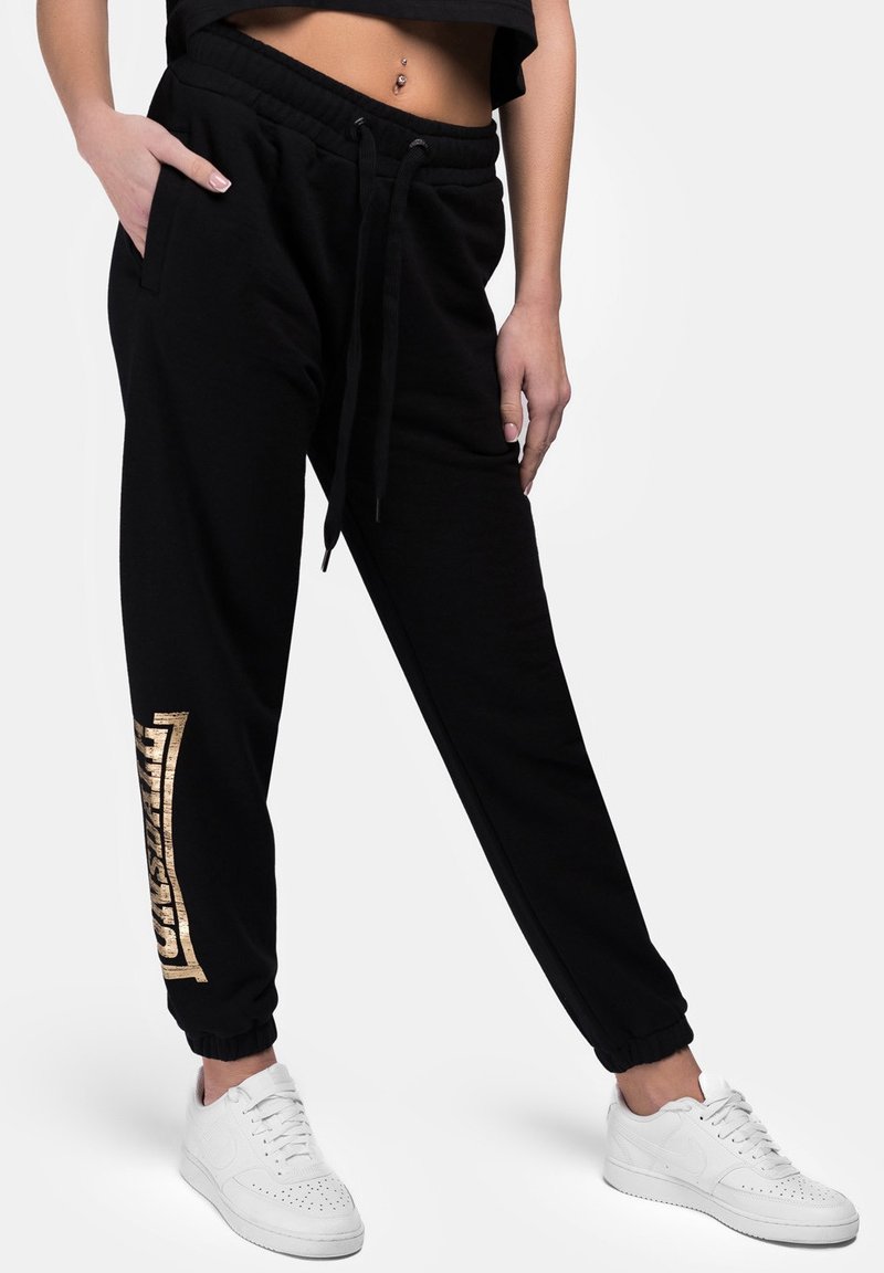 Black sweatpants with an elastic waistband, side pockets, and a gold graphic design along the left leg; cuffs are fitted.